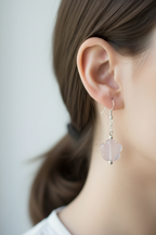 A close-up side profile of a woman's ear wearing a delicate silver dangle earring with a pale pink flower-shaped drop.