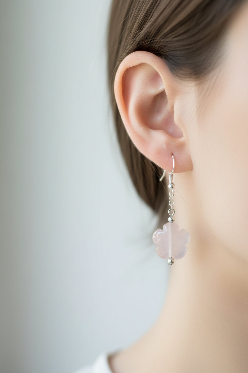 A close-up side profile of a woman's ear wearing a delicate silver dangle earring with a pale pink flower-shaped drop.