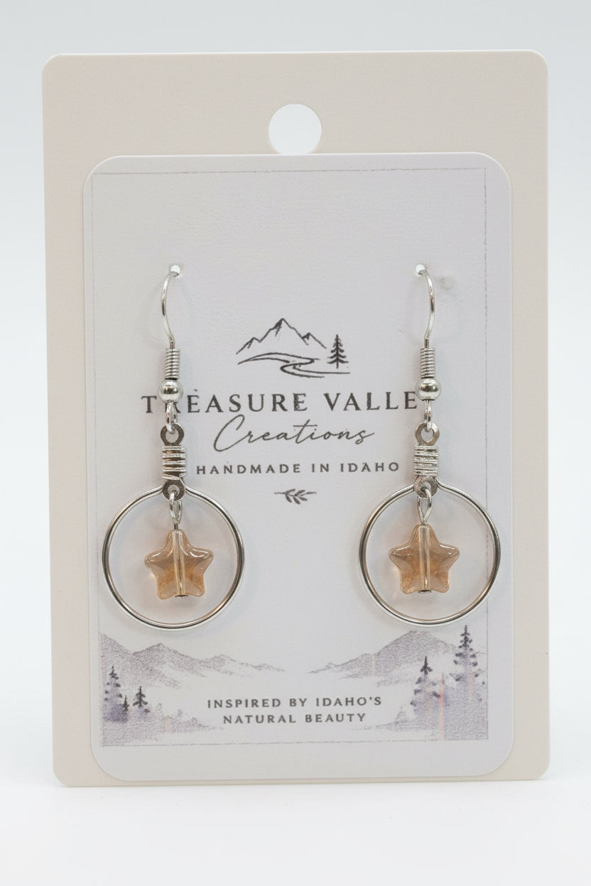 Treasure Valley Creations handmade dangle earrings with silver-toned circular hoops, wrapped wire, and star-shaped amber pendants, displayed on a branded white backing card.