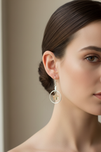 A close-up of a woman's ear and side profile wearing a delicate silver hoop earring with a small gold-toned cross charm, against a soft beige background.