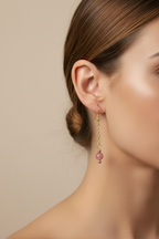 Gold dangle earrings with a small pink bead at the bottom, displayed against a black background.