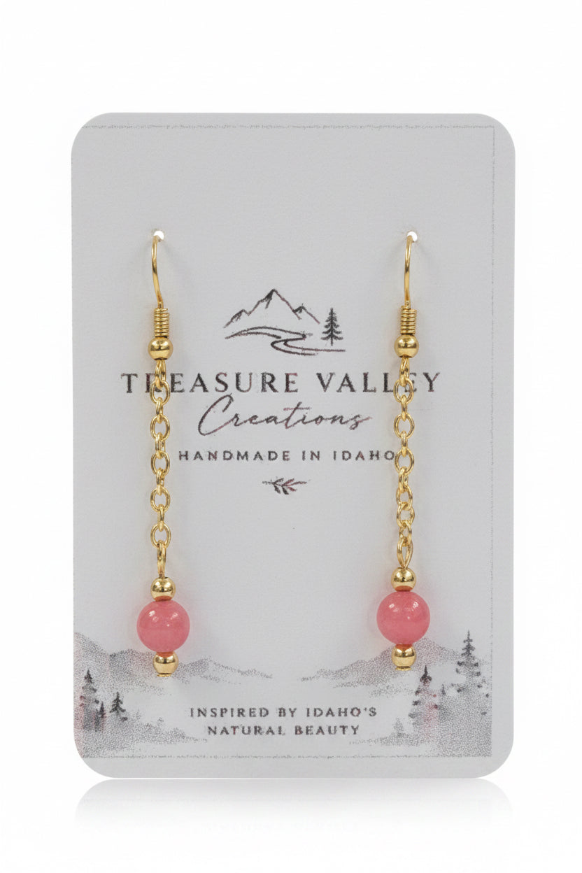A pair of gold dangle earrings with pink round beads, displayed on a white backing card from Treasure Valley Creations.