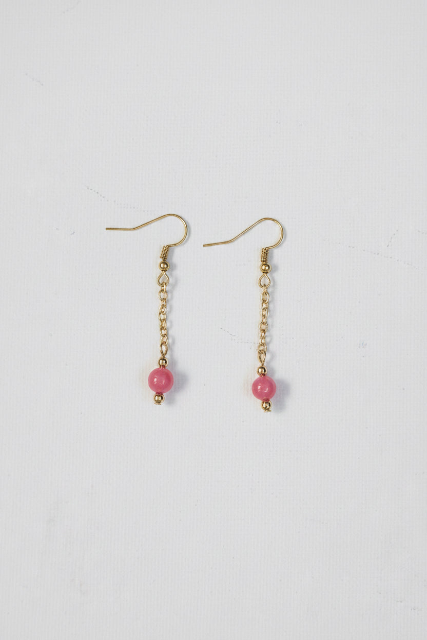 A pair of gold dangle earrings with small pink beads, displayed on a textured off-white background.
