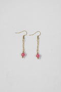 A pair of gold dangle earrings with small pink beads, displayed on a textured off-white background.