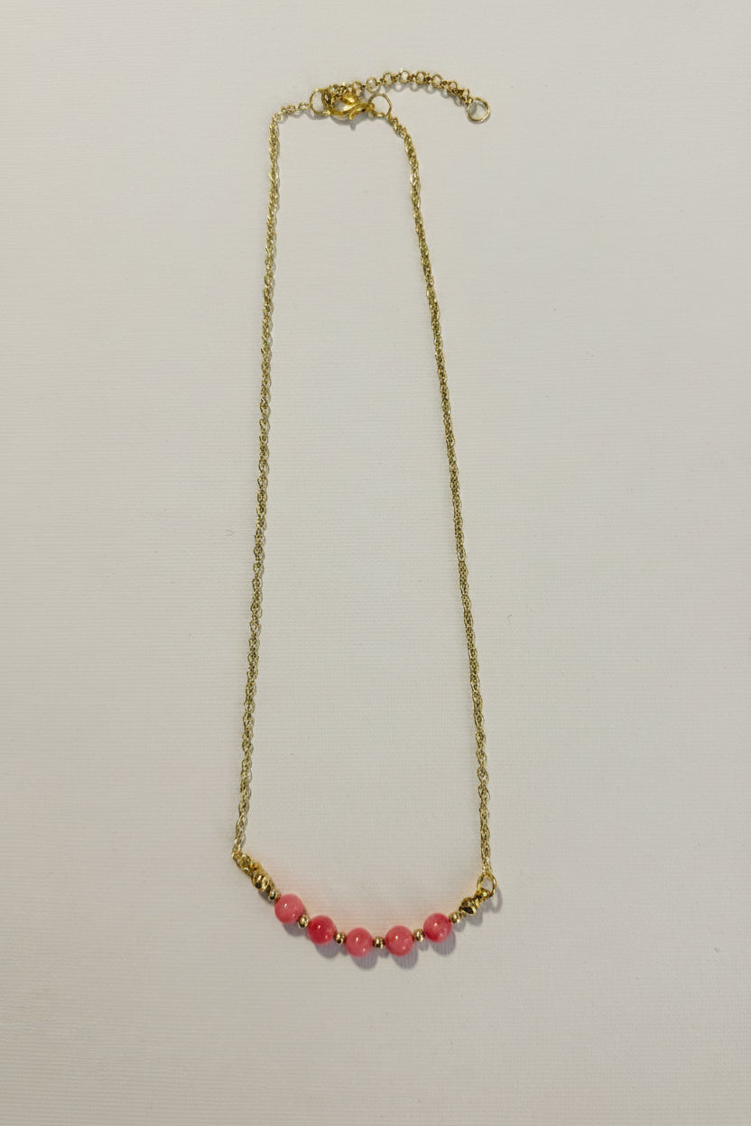 A delicate gold chain necklace featuring a row of small, round pink beads at the center, displayed against a plain off-white background.