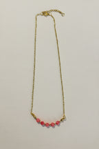 A delicate gold chain necklace featuring a row of small, round pink beads at the center, displayed against a plain off-white background.
