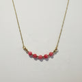 A delicate gold chain necklace featuring five smooth, round pinkish-red beads spaced along the chain, displayed on a textured off-white surface.