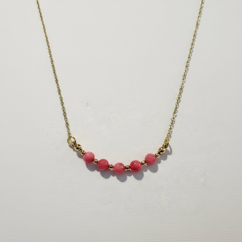 A delicate gold chain necklace featuring five smooth, round pinkish-red beads spaced along the chain, displayed on a textured off-white surface.