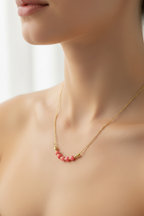 A close-up of a model's neck and upper chest wearing a delicate gold chain necklace with a curved row of small pink beads at the center.