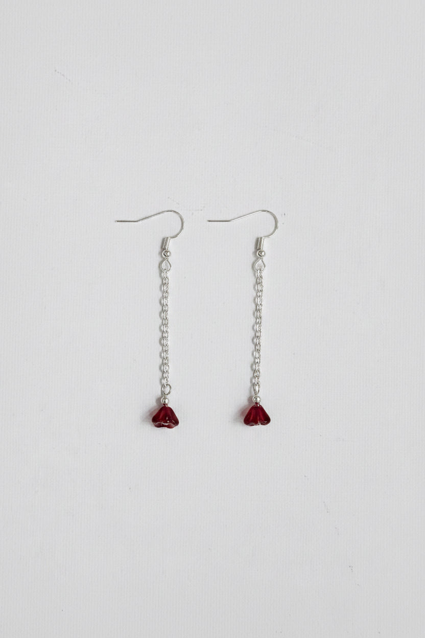 A pair of silver dangle earrings with small chain links and faceted dark red heart-shaped crystal pendants, displayed on a textured off-white fabric background.