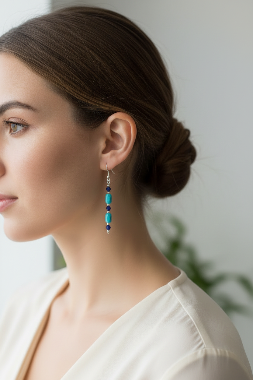 A close-up of a woman wearing a long, dangling beaded earring with turquoise blue, dark blue, and silver-toned beads, paired with a cream-colored top.