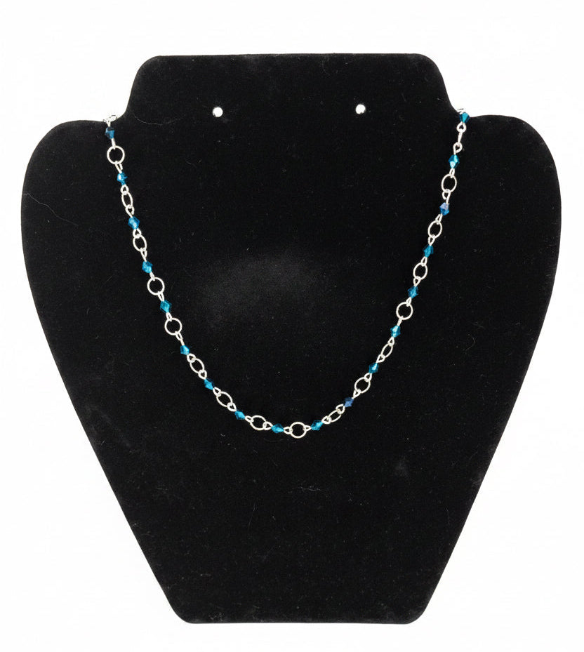 A silver chain necklace with small blue bead accents, displayed on a black jewelry bust against a white background.