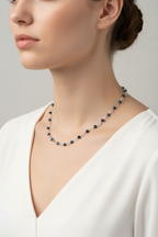 A close-up of a woman wearing a delicate beaded necklace with dark blue and clear faceted beads, paired with a white V-neck top against a neutral gray background.