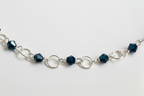 A silver chain bracelet featuring faceted dark teal beads and openwork circular links, displayed against a plain light gray background.