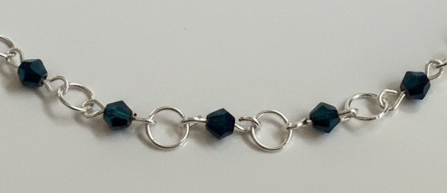 A silver chain bracelet featuring faceted dark teal beads and openwork circular links, displayed against a plain light gray background.
