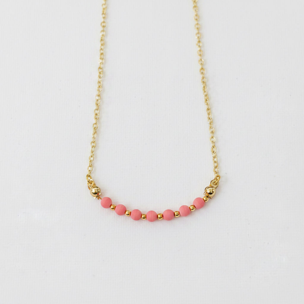 A delicate gold chain necklace featuring a curved row of small pink beads separated by tiny gold spacer beads, displayed on a textured light gray background.