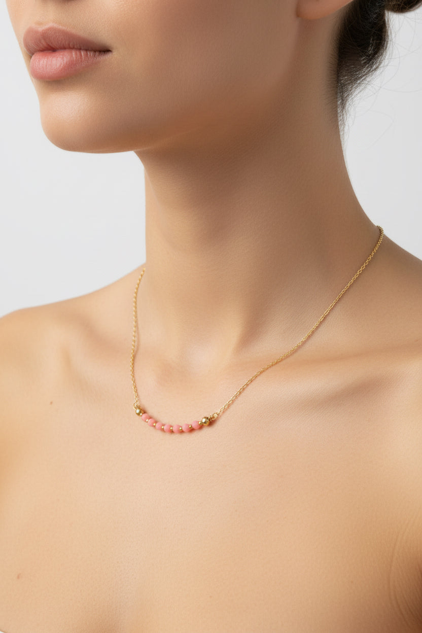 A close-up of a model's neck and upper chest wearing a delicate gold chain necklace with a curved bar of small pink beads at the center.