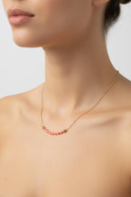 A close-up of a model's neck and upper chest wearing a delicate gold chain necklace with a curved bar of small pink beads at the center.