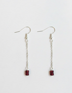A pair of silver dangle earrings with small rectangular dark red gemstone charms, displayed on a clean white background.