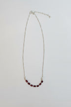 A delicate silver chain necklace featuring a curved section of small, dark red beads, displayed on a plain light gray background.