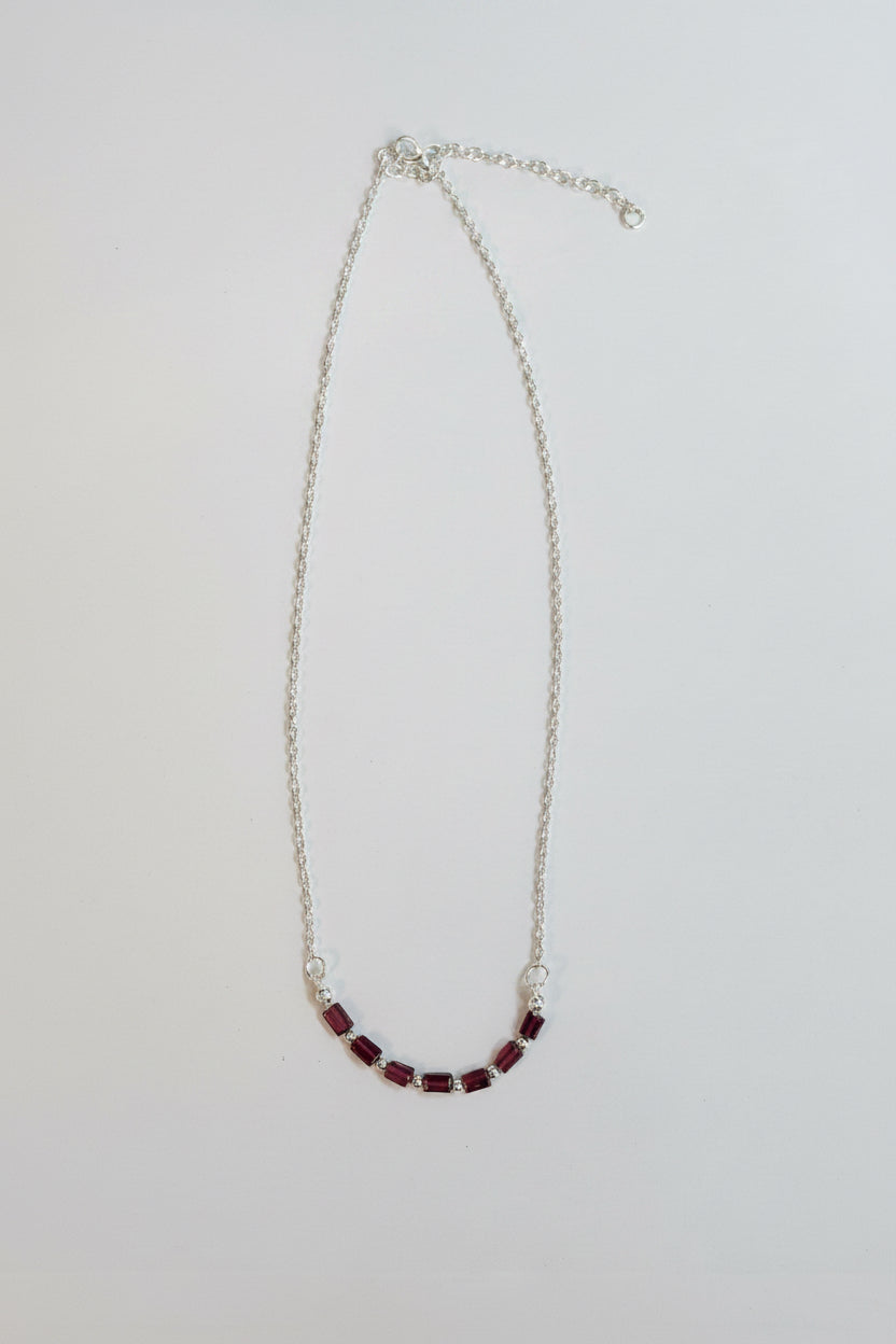 A delicate silver chain necklace featuring a curved section of small, dark red beads, displayed on a plain light gray background.