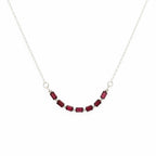 A delicate silver chain necklace featuring a curved row of small rectangular dark red gemstones, displayed on a plain light gray background.