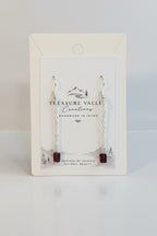 Treasure Valley jewelry display card holding a pair of silver-toned drop earrings with small chain links and dark red bead ends, photographed on a light surface.