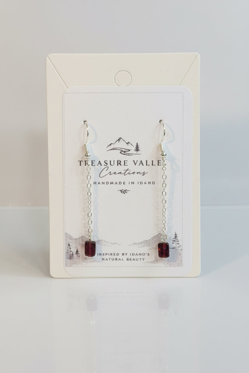 Treasure Valley jewelry display card holding a pair of silver-toned drop earrings with small chain links and dark red bead ends, photographed on a light surface.