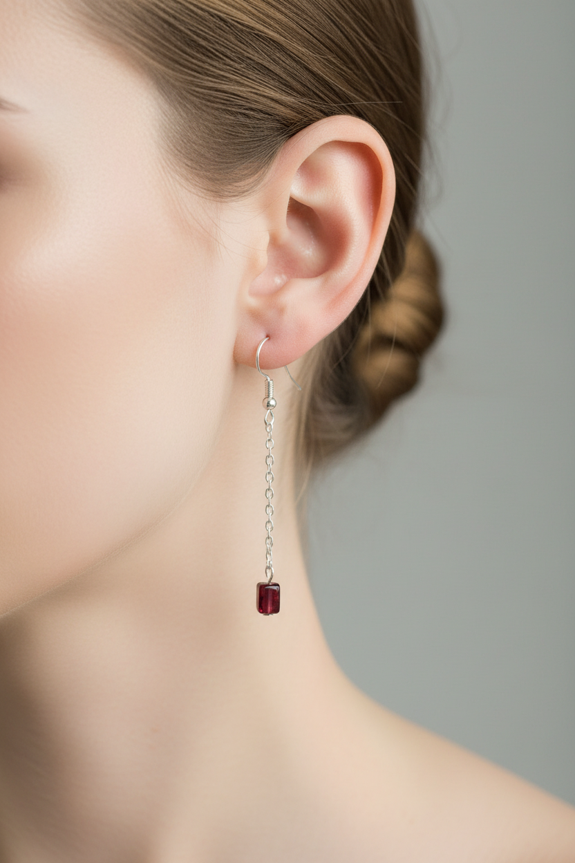 A close-up of a woman's ear wearing a silver dangle earring with a small dark red rectangular drop, against a soft gray background.