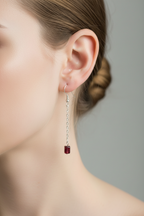 A close-up of a woman's ear wearing a silver dangle earring with a small dark red rectangular drop, against a soft gray background.