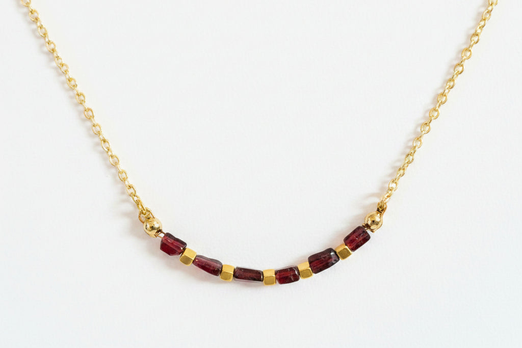 A delicate gold chain necklace featuring a row of faceted dark red beads separated by small gold spacer beads, displayed on a textured white surface.