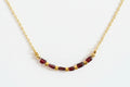 A delicate gold chain necklace featuring a row of faceted dark red beads separated by small gold spacer beads, displayed on a textured white surface.
