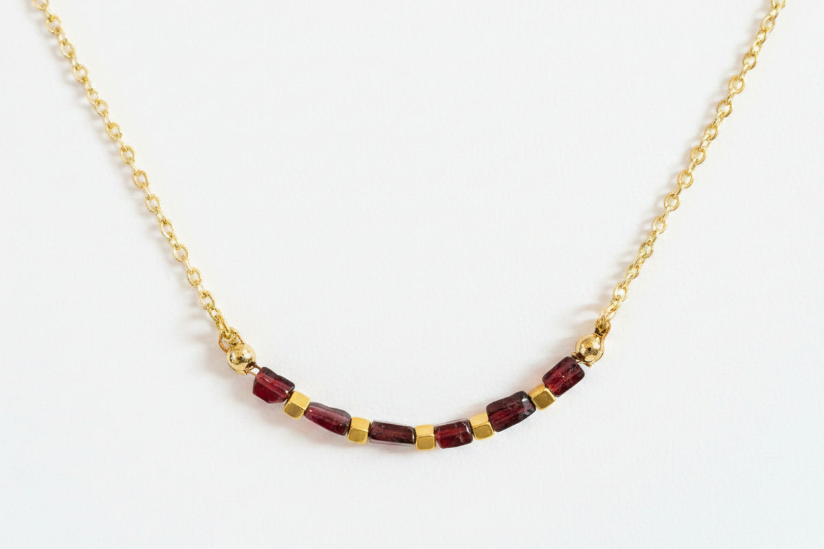 A delicate gold chain necklace featuring a row of faceted dark red beads separated by small gold spacer beads, displayed on a textured white surface.