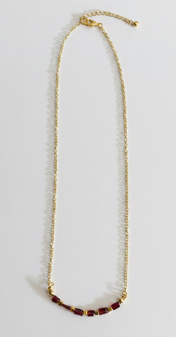 A gold chain necklace with a small cluster of dark red beads at the bottom, displayed on a plain light gray surface.