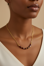 A close-up of a model wearing a delicate gold chain necklace with small rectangular dark red and gold beads at the center, styled with a white top against a warm beige background.