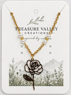 Treasure Valley Creations necklace featuring a gold chain with white pearl beads and a black-and-white rose pendant, displayed on a branded white card with floral and mountain illustrations.