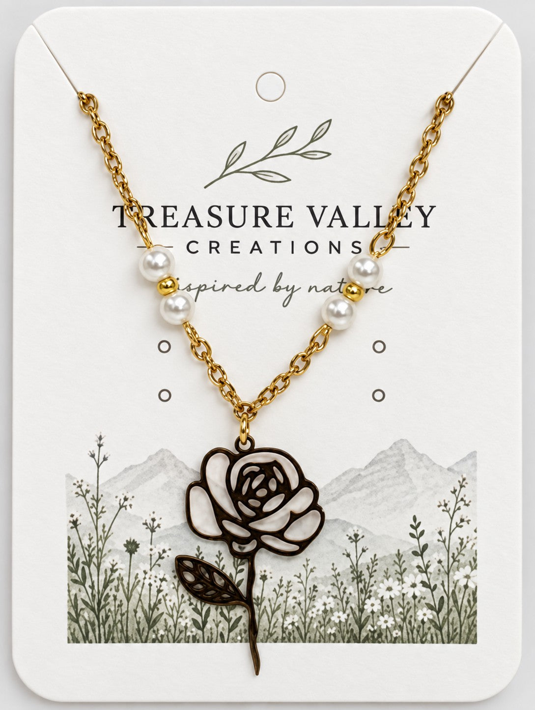 Treasure Valley Creations necklace featuring a gold chain with white pearl beads and a black-and-white rose pendant, displayed on a branded white card with floral and mountain illustrations.