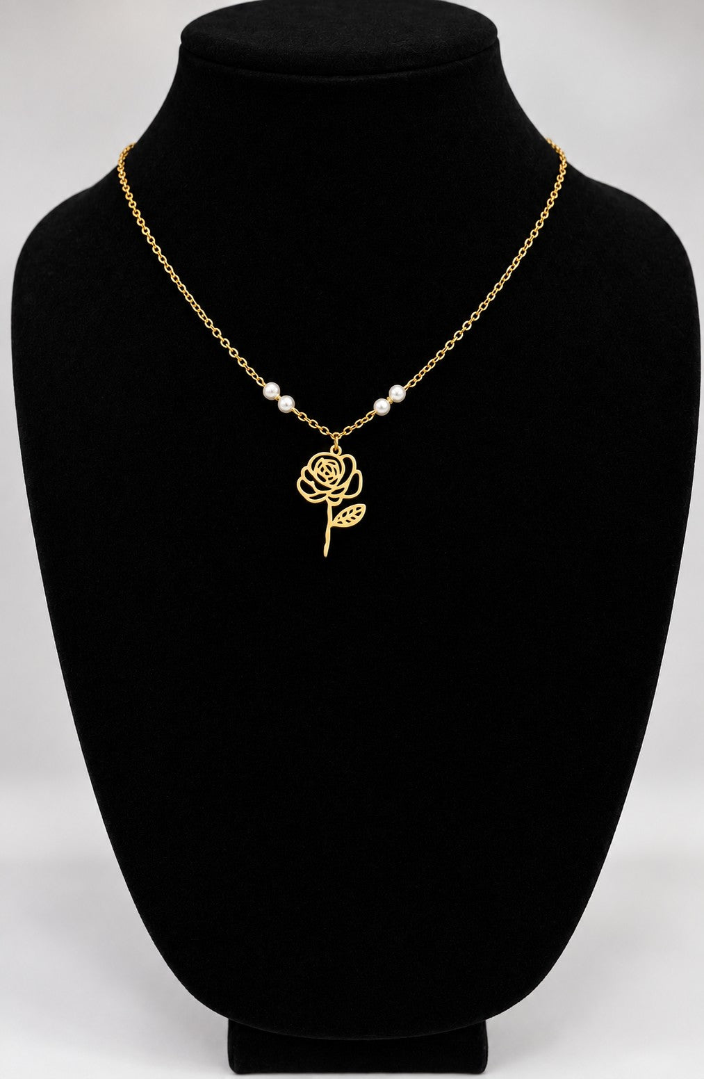 A gold chain necklace with a rose-shaped pendant and three small pearl-like beads, displayed on a black jewelry bust against a light gray background.