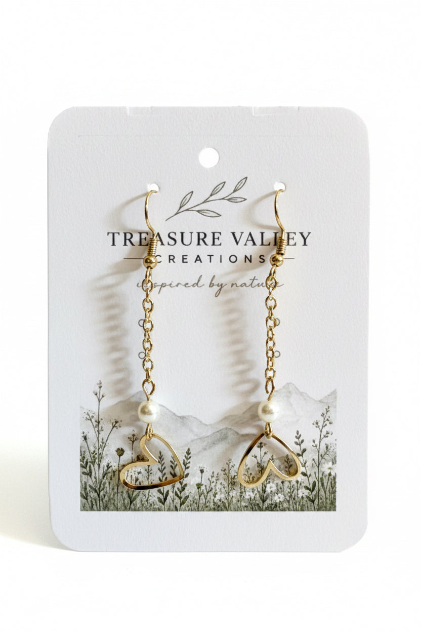 A pair of gold dangle earrings with open heart charms and small pearl beads, displayed on a white jewelry card from Treasure Valley Creations.
