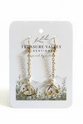 A pair of gold dangle earrings with open heart charms and small pearl beads, displayed on a white jewelry card from Treasure Valley Creations.