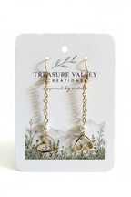 A pair of gold dangle earrings with open heart charms and small pearl beads, displayed on a white jewelry card from Treasure Valley Creations.