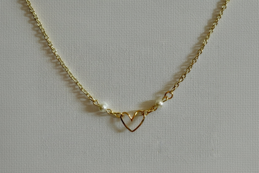 A gold chain necklace featuring a central open heart pendant and two small white pearl beads on either side, displayed on a textured light gray fabric background.