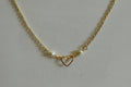 A gold chain necklace featuring a central open heart pendant and two small white pearl beads on either side, displayed on a textured light gray fabric background.