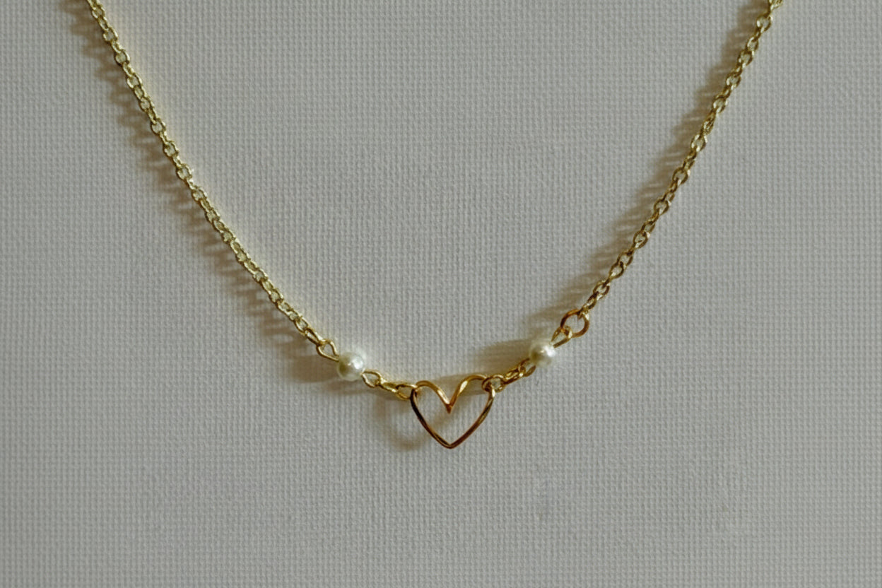 A gold chain necklace featuring a central open heart pendant and two small white pearl beads on either side, displayed on a textured light gray fabric background.