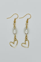 A pair of gold dangle earrings featuring a freshwater pearl and an open heart-shaped gold pendant, displayed on a neutral light gray background.