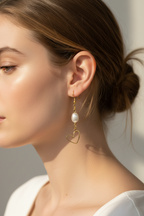 A close-up side profile of a woman wearing a delicate gold dangle earring with a white pearl and a small open heart charm, set against a soft neutral background.