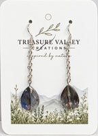 A pair of long dangle earrings with silver-toned chains and faceted dark blue-gray teardrop crystals, displayed on a white backing card printed with the brand name Treasure Valley Creations.