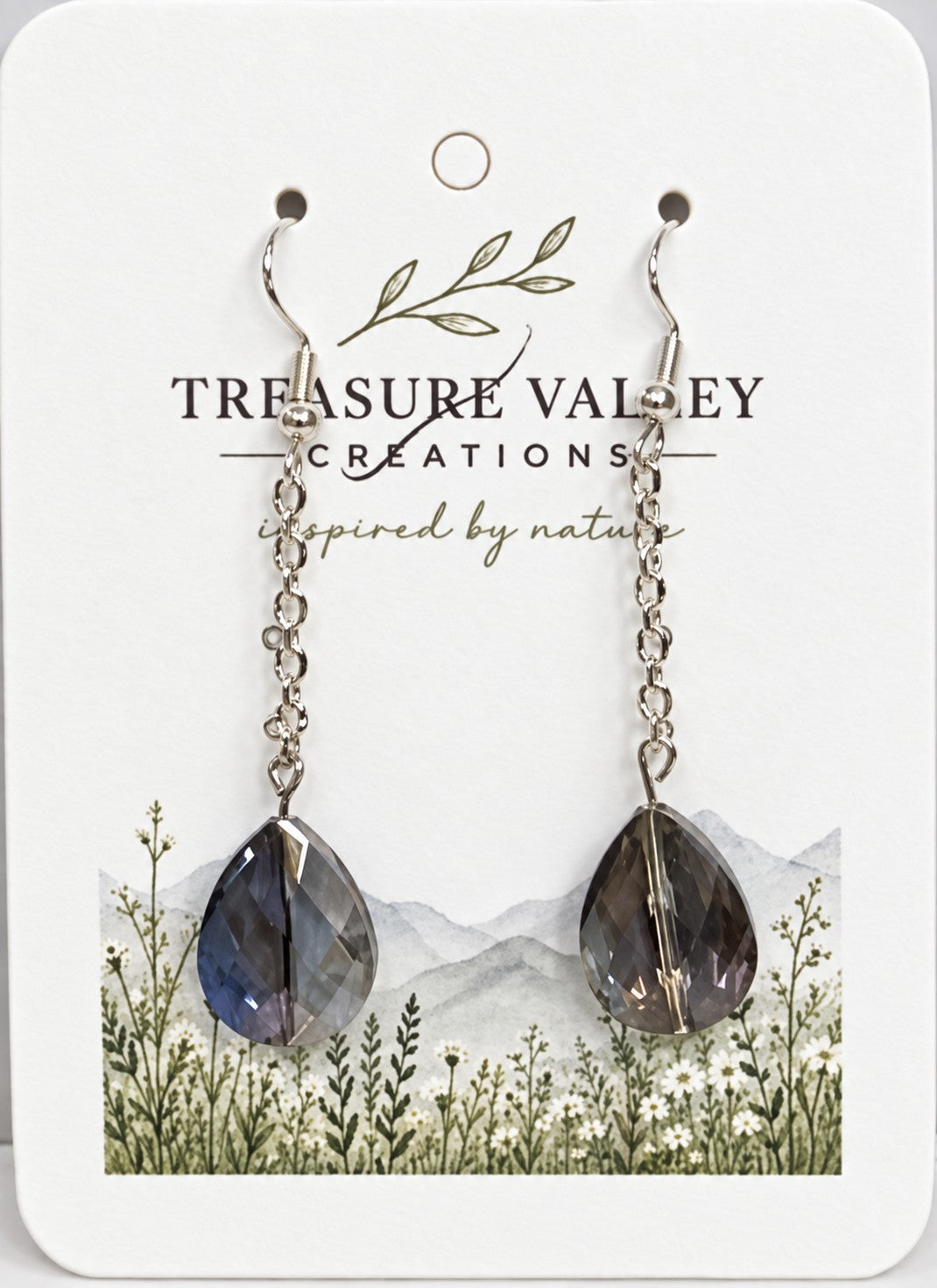 A pair of long dangle earrings with silver-toned chains and faceted dark blue-gray teardrop crystals, displayed on a white backing card printed with the brand name Treasure Valley Creations.