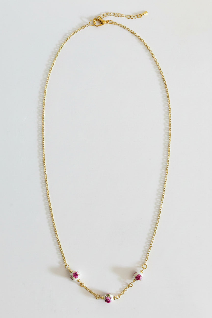 A delicate gold chain necklace with three small pink and white floral bead accents at the bottom center, displayed on a plain light gray background.