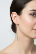 A close-up of a woman's ear and side profile wearing a long silver dangle earring with small green and clear faceted beads, against a light gray background.